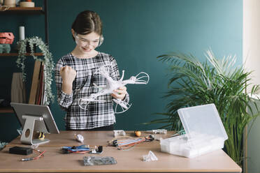 Smiling girl holding drone gesturing while standing against wall at home - ALBF01527