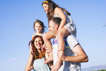 Cheerful parents carrying daughters on shoulders against clear sky during sunny day - JCMF01349