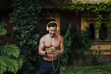 Shirtless mid adult man listening music through headphones while standing in yard - EBBF00700