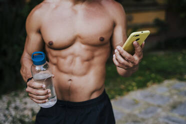 Close-up of shirtless male athlete holding water bottle using smart phone in yard - EBBF00701