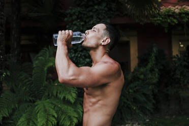Hemdloser männlicher Sportler, der Wasser trinkt, während er gegen Pflanzen im Hof steht - EBBF00702