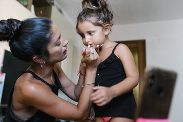 Mother putting lipstick to daughter while video recording at home - EGAF00697