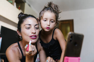 Mother and daughter applying lipstick while video recording in smart phone at home - EGAF00698
