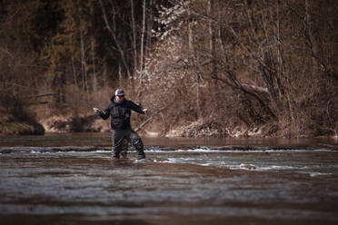 Fly fisherman holding fishing rod while casting in flowing river at forest - DHEF00382