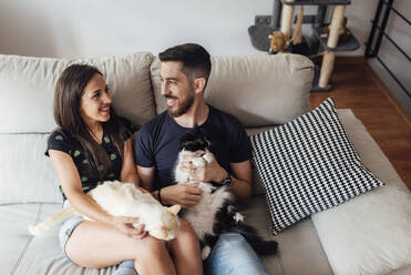 Smiling couple sitting with cats on sofa at home - SASF00060
