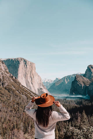 Back view of unrecognizable female traveler in hat standing on hill covered with forest and admiring picturesque scenery with granite cliffs in sunny day in Yosemite National Park in California stock photo