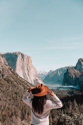 Back view of unrecognizable female traveler in hat standing on hill covered with forest and admiring picturesque scenery with granite cliffs in sunny day in Yosemite National Park in California - ADSF15243