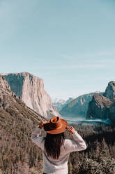 Rückenansicht einer nicht erkennbaren weiblichen Reisenden mit Hut, die auf einem mit Wald bedeckten Hügel steht und die malerische Landschaft mit Granitfelsen an einem sonnigen Tag im Yosemite-Nationalpark in Kalifornien bewundert - ADSF15243