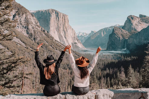 Zurück Blick auf glückliche unerkennbar weibliche Freunde in trendigen Kleidern und Hüte sitzen am Rande der felsigen Klippe und genießen erstaunliche Berglandschaft beim Besuch Yosemite National Park in Kalifornien in sonnigen Frühlingstag - ADSF15245