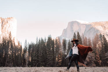 Seitenansicht einer fröhlichen jungen Frau in Poncho und Hut, die auf trockenem Gras in der Nähe des Waldes mit Granitfelsen im Hintergrund an einem sonnigen Tag im Yosemite National Park in den USA spazieren geht - ADSF15246
