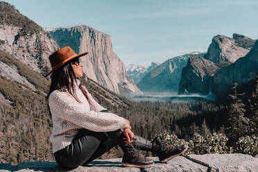 Ganzkörper-Seitenansicht von glücklichen entspannten jungen weiblichen Reisenden in stilvollem Outfit sitzen auf Stein Grenze gegen malerische Berglandschaft mit felsigen Klippen und Nadelwald in Yosemite National Park in USA - ADSF15247