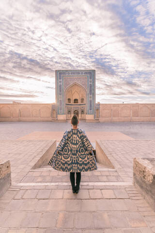 Back view of female in ornamental coat standing on aged square against cloudy sky in Bukhara, Uzbekistan stock photo