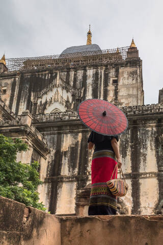 From below back view of woman in long dress holding traditional Burmese red umbrella and bag standing next to old stone building while walking and sightseeing in Bagan Myanmar stock photo