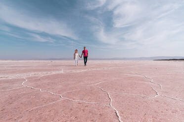 From below faceless couple in vivid casual clothes looking away and enjoying view while holding hands and walking along pink dry shore of amazing salt lake under blue sky with white lush clouds - ADSF15270