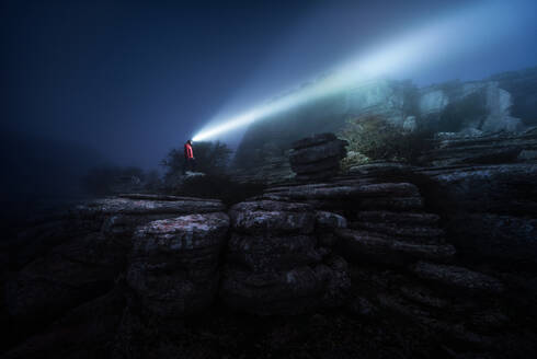 Seitenansicht eines Touristen, der auf einer felsigen Klippe steht und raue Felsbrocken in einer nebligen Nacht beleuchtet - ADSF15310