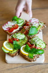 Crop female with delicious toasts with cucumbers and tomatoes garnished with fresh lemon and sprigs of basil and placed on wooden chopping board - ADSF15324