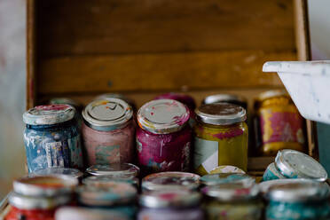 Plastikflaschen mit bunten Farben und leeren Behältern in der Nähe von Künstlerwerkzeugen auf schmutzigen Tisch in der Nähe von bunten Wand mit unebener Oberfläche in Kunststudio - ADSF15344