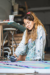 Fröhliche barfüßige Künstlerin in schmutzigem Gewand und Handschuhen, die mit geschlossenen Augen vor einem abstrakten Gemälde im Kunststudio auf dem Holzboden liegt - ADSF15345