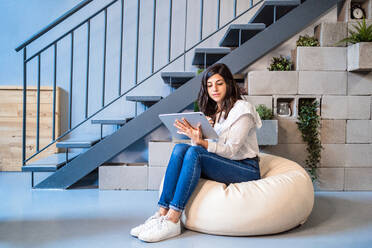 Full length of focused young female designer in casual clothes sitting on comfortable pouf and using graphic tablet while working in contemporary creative workspace - ADSF15349