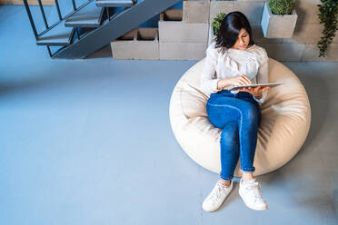 Top view full length of focused young female designer in casual clothes sitting on comfortable pouf and using graphic tablet while working in contemporary creative workspace - ADSF15350