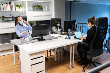 Serious young man in medical mask discussing business issue by phone while female colleagues working with computers in contemporary coworking office - ADSF15353