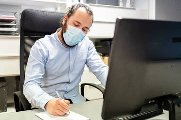 Fokussierter junger bärtiger männlicher Manager in formellem blauem Hemd und Schutzmaske macht sich Notizen in einem Notizbuch, während er auf einem Stuhl vor einem Computerbildschirm an einem modernen Arbeitsplatz sitzt - ADSF15355