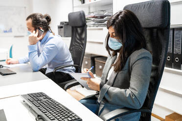 Serious young female employee in formal clothes and protective mask sitting at desk with computer and writing in notebook while working in contemporary workspace with colleague - ADSF15356