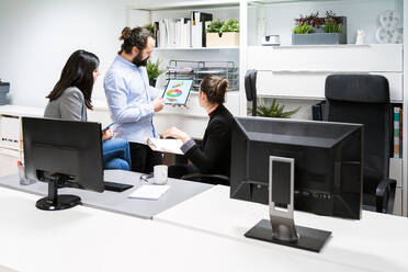 Young male manager holding tablet with diagrams and analyzing financial results of business project during meeting with female coworkers in modern workspace - ADSF15359