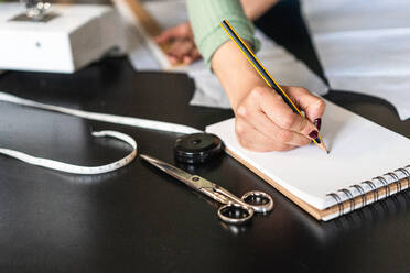 Junge ethnische Näherin mit Brille macht sich Notizen, während sie nach vorne gelehnt steht und ein Lineal an einem Tisch mit Papieren und Scheren in einem Atelier im Loft-Stil benutzt - ADSF15365