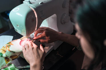 Von oben Ernte unerkennbar Handwerkerin mit modernen Nähmaschine bei der Herstellung von weichen Maske mit kreativen grünen Muster in der Nähe von Lampe in Loft-Stil Workshop - ADSF15378