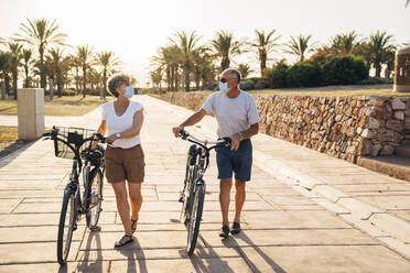 Senior man and woman with face mask talking while walking with cycle at park - MPPF01083