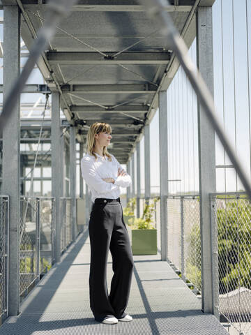 Thoughtful businesswoman with arms crossed standing on footbridge in greenhouse stock photo