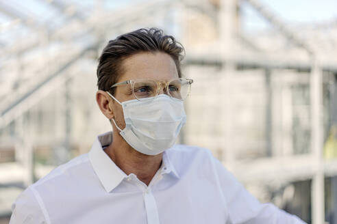 Close-up of businessman wearing face mask looking away in greenhouse - JOSEF01638