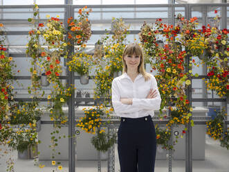 Smiling businesswoman with arms crossed standing against flowers in greenhouse - JOSEF01643