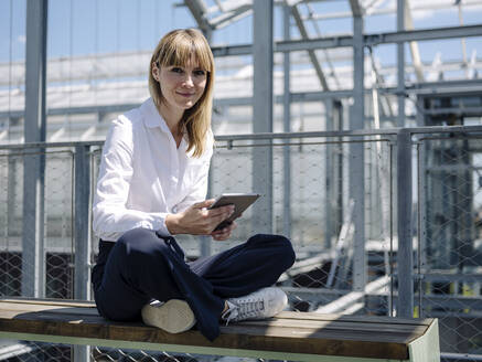 Lächelnde Geschäftsfrau, die ein digitales Tablet benutzt, während sie auf einem Sitz im Gewächshaus sitzt - JOSEF01656