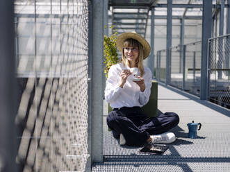 Geschäftsfrau mit Hut und Kaffeetasse am Zaun im Gewächshaus sitzend - JOSEF01658