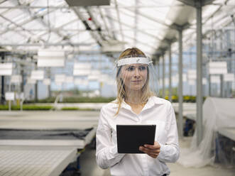 Businesswoman wearing face shield with digital tablet working in plant nursery - JOSEF01663