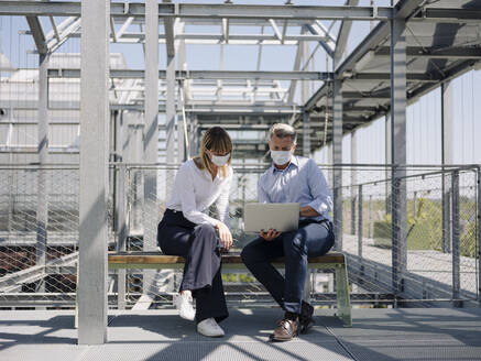 Colleagues wearing masks discussing over laptop while sitting on seat in greenhouse - JOSEF01678