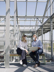 Business people wearing masks discussing over laptop while sitting on seat in greenhouse - JOSEF01679