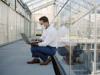 Businessman wearing mask using laptop while crouching on floor in greenhouse - JOSEF01685