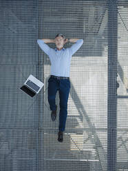 Businessman with hands behind head lying on metallic floor in greenhouse - JOSEF01687