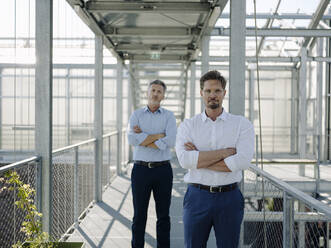 Confident male coworkers with arms crossed standing on footbridge in greenhouse - JOSEF01695