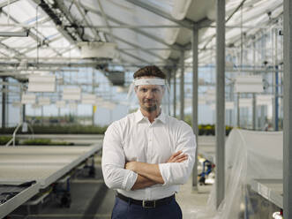 Businessman with arms crossed wearing face shield while standing in greenhouse - JOSEF01697