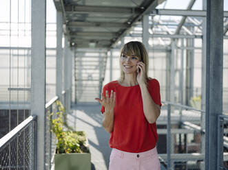 Smiling businesswoman talking over smart phone while standing on footbridge in greenhouse - JOSEF01703