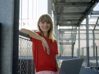 Smiling businesswoman using laptop while sitting by railing in greenhouse - JOSEF01704