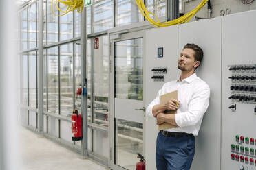 Nachdenklicher Geschäftsmann hält ein Klemmbrett in der Hand und lehnt sich an eine Schalttafel im Gewächshaus - JOSEF01727