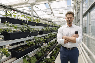 Confident male entrepreneur with arms crossed standing by plants in greenhouse - JOSEF01736