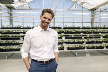 Smiling male entrepreneur with hands in pockets standing against plants at greenhouse - JOSEF01739