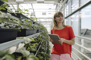 Lächelnde Unternehmerin mit digitalem Tablet, die wegschaut, während sie bei Pflanzen in einer Gärtnerei steht - JOSEF01771