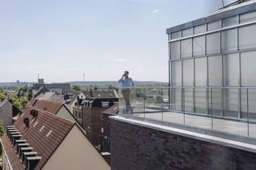 Männlicher Berufstätiger, der über sein Smartphone spricht, während er auf einem Balkon gegen den Himmel steht - JOSEF01805
